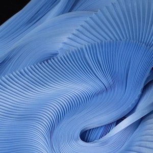 Op de afbeelding: Abstracte afbeelding van lichtblauwe geplooide stof. De stof is gerangschikt in vloeiende, golfachtige patronen, wat een gevoel van beweging en textuur creëert. De plooien zijn dicht op elkaar geplaatst, wat diepte en dimensie toevoegt.