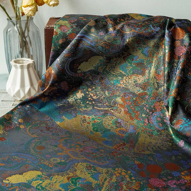 Puede incluir: Una tela drapeada con un patr&oacute;n rico e intrincado. El dise&ntilde;o presenta una mezcla de colores que incluyen verde azulado, dorado, naranja y azules profundos. La superficie de la tela tiene una calidad brillante, lo que sugiere un material de sat&eacute;n o seda. La est&eacute;tica general es lujosa y art&iacute;stica.