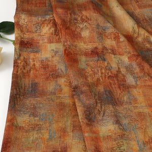 May include: Close-up of a fabric with a textured, abstract design. The fabric features a blend of warm colors, including orange, brown, and yellow, with hints of blue. The pattern appears to be a series of overlapping rectangular shapes.