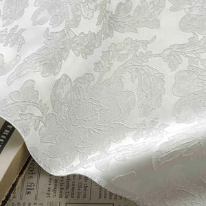 May include: Close-up of a white fabric with a raised floral pattern. The design features detailed, embossed flowers and leaves, creating a textured surface. The fabric has a slight sheen, enhancing the intricate details of the pattern.