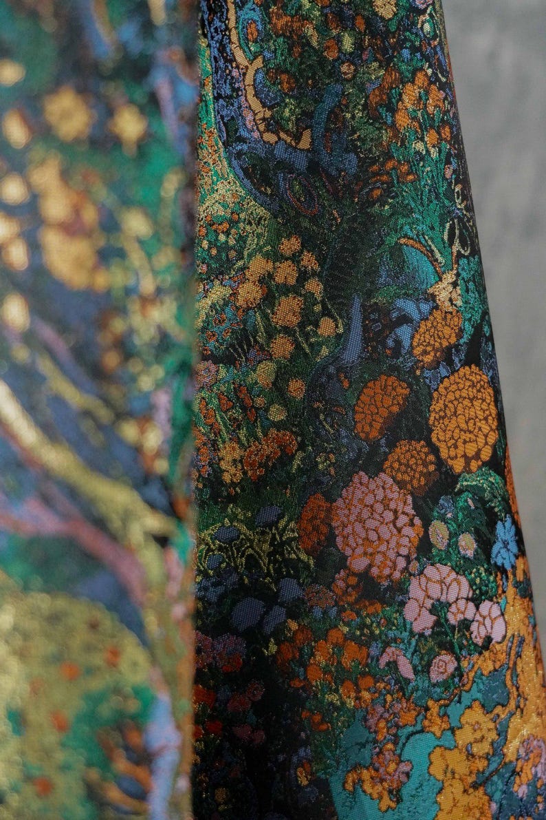 Puede incluir: Primer plano de una tela con un estampado floral detallado. El dise&ntilde;o presenta un fondo oscuro con vibrantes flores naranjas, rosas y verdes, junto con acentos dorados. La tela tiene una apariencia lujosa y texturizada.