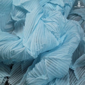 May include: Close-up of light blue pleated fabric. The fabric has a textured, rippled appearance with vertical and diagonal folds. The material is bunched and layered, creating shadows and depth. The image is well-lit, highlighting the fabric's texture.