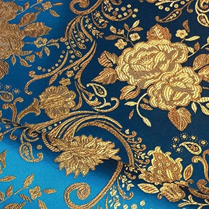 Pode incluir: Tecido azul com um padrão floral dourado. O tecido tem uma textura de brocado e apresenta um desenho repetitivo de flores e folhas.