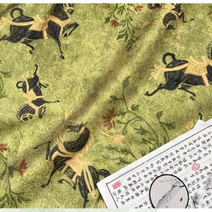 Może przedstawiać: Zielona tkanina z powtarzającym się wzorem czarnych koni z jeźdźcami i kwiatowymi akcentami. Kawałek papieru z azjatyckimi znakami znajduje się w prawym dolnym rogu.