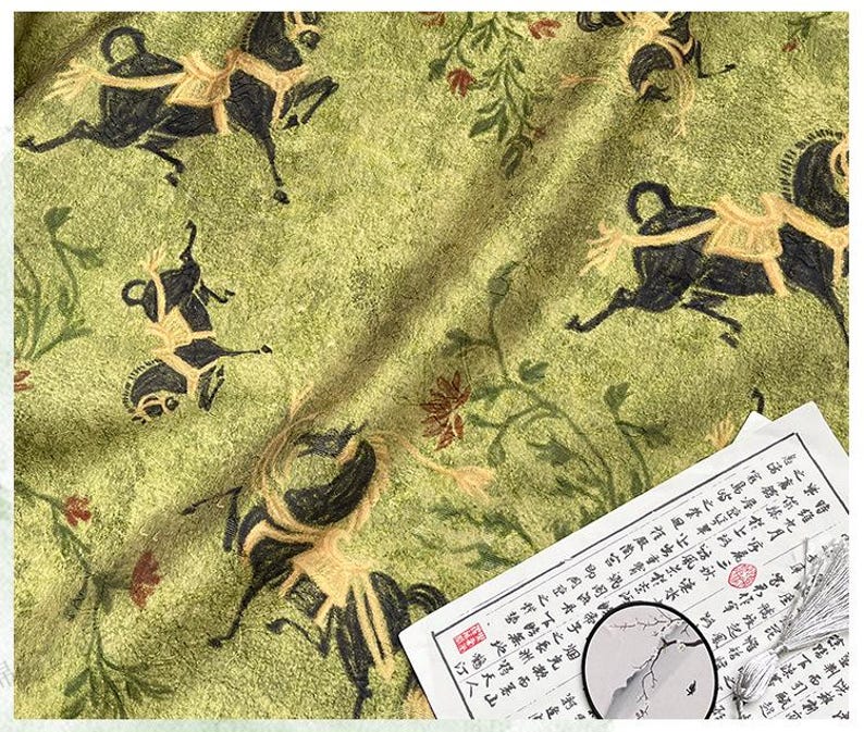 Może przedstawiać: Zielona tkanina z powtarzającym się wzorem czarnych koni z jeźdźcami i kwiatowymi akcentami. Kawałek papieru z azjatyckimi znakami znajduje się w prawym dolnym rogu.