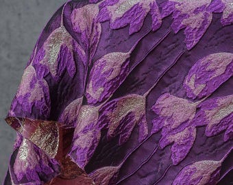Può includere: Primo piano di un lussuoso tessuto viola con un motivo floreale. Il tessuto presenta forme di foglie stilizzate in rilievo in tonalità viola e argento, creando un effetto strutturato. Il tessuto è piegato, rivelando uno scorcio di una fodera dorata a contrasto.