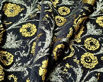 May include: Close-up of a dark fabric with a repeating floral pattern. The design features yellow flowers and silver-gray foliage against a black background. The fabric has a textured appearance with folds and shadows.