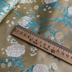 Può includere: Primo piano di un tessuto dorato con un motivo floreale bianco e turchese. Un righello di legno con il testo "Made in China" è posizionato sopra il tessuto.