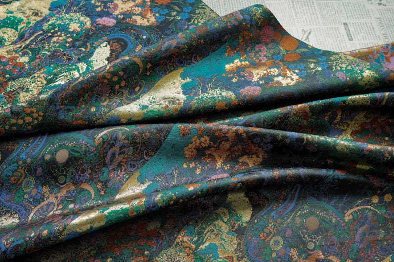 Puede incluir: Una tela doblada con un patr&oacute;n detallado y colorido. El dise&ntilde;o presenta una mezcla de tonos turquesa, dorado, marr&oacute;n y azul, con motivos florales y tipo paisley. La tela tiene un brillo lujoso, lo que sugiere un material de alta calidad.