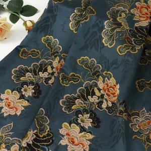 Könnte beinhalten: Nahaufnahme eines teal-farbenen Stoffes mit einem gestickten Blumenmuster. Das Design zeigt große Blüten in Orange, Creme und Blau, mit grünen und goldenen Blättern. Der Stoff hat einen subtilen Glanz und ein strukturiertes Aussehen, was auf ein hochwertiges Material hindeutet.