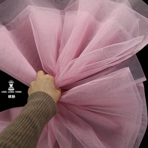Può includere: Un primo piano di un tessuto di tulle rosa pallido. Il tessuto è morbido e delicato, con una finitura leggermente scintillante. Il tessuto è raccolto e drappeggiato, creando un aspetto fluido ed etereo.
