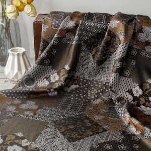 Può includere: Un tessuto patchwork con una varietà di motivi nei toni del marrone, oro e bianco. Il tessuto presenta disegni floreali, geometrici e astratti. Il tessuto è drappeggiato, creando pieghe e ombre.