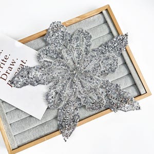 May include: A large, silver sequined flower applique with intricate detailing. The applique is on a gray velvet surface within a wooden frame. A white paper with the words "Write. Draw. Repeat." is partially visible.
