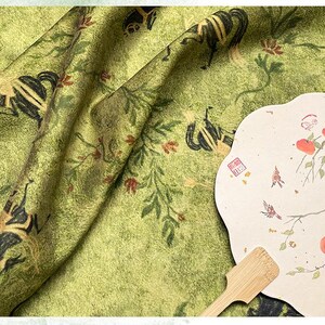 Może przedstawiać: Zbliżenie zielonej tkaniny z powtarzającym się wzorem czarnych i złotych postaci, liści i kwiat&oacute;w. Po prawej stronie papierowy wachlarz z akwarelą ptak&oacute;w i owoc&oacute;w. U dołu drewniany uchwyt.