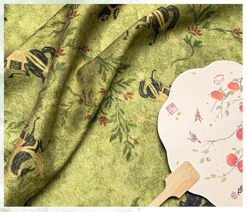 Może przedstawiać: Zbliżenie zielonej tkaniny z powtarzającym się wzorem czarnych i złotych postaci, liści i kwiat&oacute;w. Po prawej stronie papierowy wachlarz z akwarelą ptak&oacute;w i owoc&oacute;w. U dołu drewniany uchwyt.