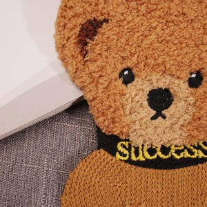 Pode incluir: Pelúcia de urso marrom vestindo um suéter marrom com detalhes pretos e a palavra "Success" bordada em amarelo.
