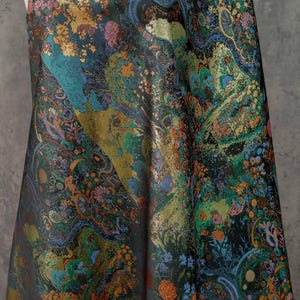 Puede incluir: Tela oscura con un intrincado estampado floral. El dise&ntilde;o presenta tonos dorados, turquesa, naranja y azul, creando una apariencia rica y texturizada. La tela drapeada muestra el estampado detallado.