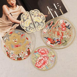 Pode incluir: Cinco patches bordados com vários desenhos. Os patches são circulares e apresentam uma variedade de cores, incluindo ouro, vermelho, rosa e azul. Os desenhos incluem uma criatura mítica, flores e um padrão chinês tradicional.