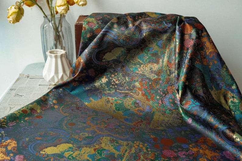 Puede incluir: Una tela drapeada con un dise&ntilde;o abstracto vibrante en tonos de verde azulado, dorado, naranja y morado. El textil tiene un brillo met&aacute;lico brillante, lo que sugiere un material lujoso. Ideal para ropa o decoraci&oacute;n del hogar.