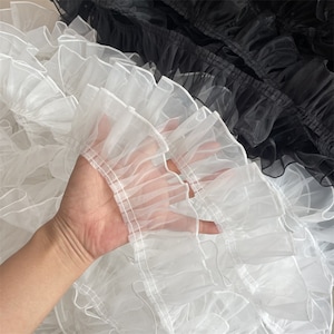 Könnte beinhalten: Weißer Rüschenbesatz mit zarter, durchsichtiger Textur. Der Besatz besteht aus mehreren Stofflagen, die einen voluminösen und eleganten Look erzeugen.