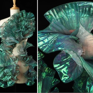 May include: A green iridescent fabric with a pleated texture. The fabric is arranged in a series of overlapping layers, creating a three-dimensional effect. The fabric is light and airy, and it has a soft, flowing drape.