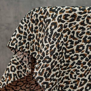Puede incluir: Primer plano de una tela con estampado de leopardo. La tela presenta un fondo color crema con manchas negras y marrones. La tela está drapeada sobre una superficie, mostrando la textura y el patrón.