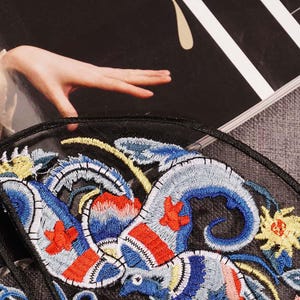 May include: A black mesh fabric patch with colorful embroidered floral and bird designs. The embroidery features shades of blue, red, yellow, and white.