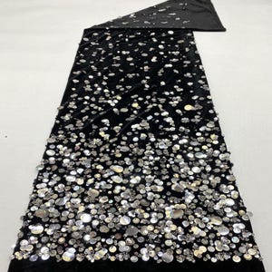 Peut inclure: Tissu noir en velours, orné de nombreuses paillettes argentées de tailles variées. Les paillettes créent un effet scintillant sur toute la surface. Le tissu est plié en haut, révélant un envers noir uni. Idéal pour les robes de soirée ou les tenues de cérémonie.