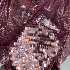 May include: Close-up of a shimmering fabric in a deep burgundy colour. The material is densely covered in small, rectangular sequins that catch the light, creating a sparkling effect. The fabric is folded, showing texture and depth.
