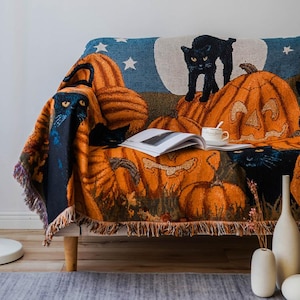 Peut inclure: Une couverture en tissu d'Halloween avec un chat noir, des citrouilles et des étoiles sur un fond bleu. La couverture est posée sur un canapé avec un livre et un vase sur la table.