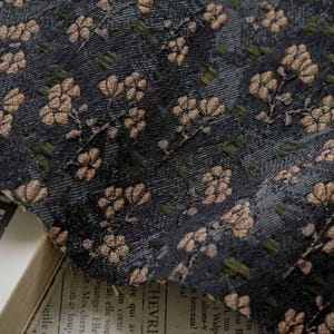 Puede incluir: Primer plano de una tela azul oscuro con un estampado floral. Las flores son en tonos beige y marrón, con pequeñas hojas verdes. La tela tiene un aspecto texturizado, y una parte de un libro es visible en la esquina inferior izquierda.