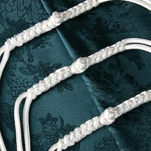 以下が含まれることがあります： 結ばれた白いサテンリボンにクリスタル装飾が施されています。リボンは濃い緑色の布地に置かれています。