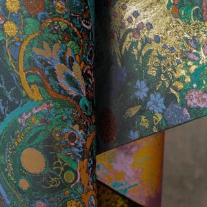 Puede incluir: Primer plano de una tela estampada con fondo oscuro. La tela presenta un dise&ntilde;o complejo con elementos florales y abstractos dorados, verdes, azules y naranjas. La tela est&aacute; doblada, revelando capas de color y textura.
