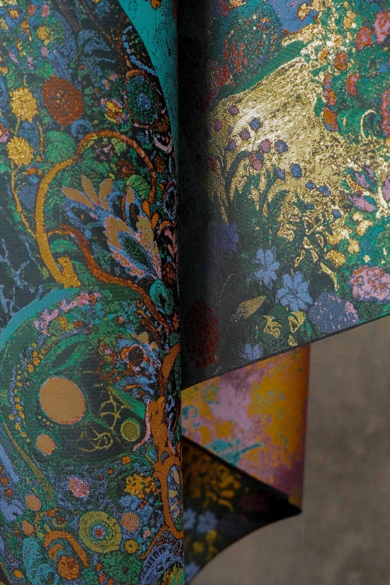 Puede incluir: Primer plano de una tela estampada con fondo oscuro. La tela presenta un dise&ntilde;o complejo con elementos florales y abstractos dorados, verdes, azules y naranjas. La tela est&aacute; doblada, revelando capas de color y textura.