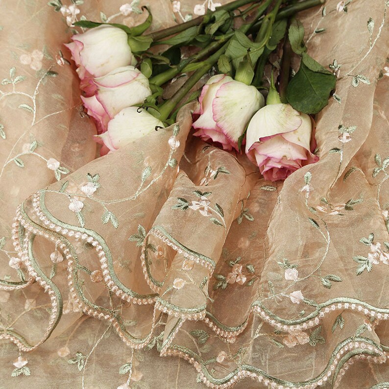 May include: A sheer peach-colored fabric with delicate floral embroidery and a scalloped edge. The fabric is adorned with a bouquet of white roses with pink edges.