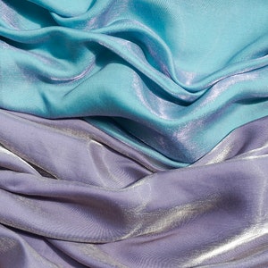 May include: Close-up of a soft, silky fabric in two shades of blue. The fabric is draped and folded, creating a textured and luxurious look.