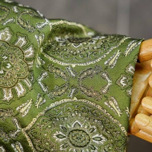 May include: A long-sleeved garment in a vibrant olive green hue, featuring an intricate, repeating woven pattern in shades of white, gold, and dark brown. The fabric displays a textured, damask-like appearance with detailed floral motifs.