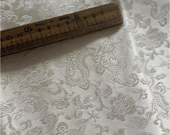 May include: Ivory-colored fabric with an embossed dragon and floral pattern. A wooden ruler, marked in inches, rests on the fabric. The fabric's design includes dragons, flowers, and swirling patterns, creating a textured surface.