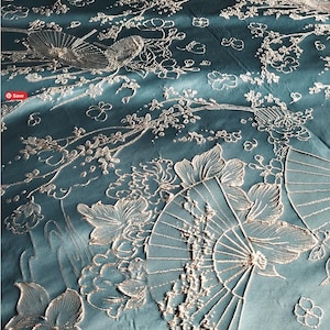 May include: Teal fabric with an embroidered floral and fan design. The embroidery is silver and features flowers, leaves, and fans. The fabric has a subtle sheen, creating a luxurious appearance. The design is intricate and detailed, suitable for home decor or apparel.