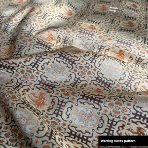 May include: Close-up of a patterned fabric with a beige background. The fabric features a repeating design of brown, orange, and white shapes. The text "Warring states pattern" is visible. The text "Multiple strings" is also visible.