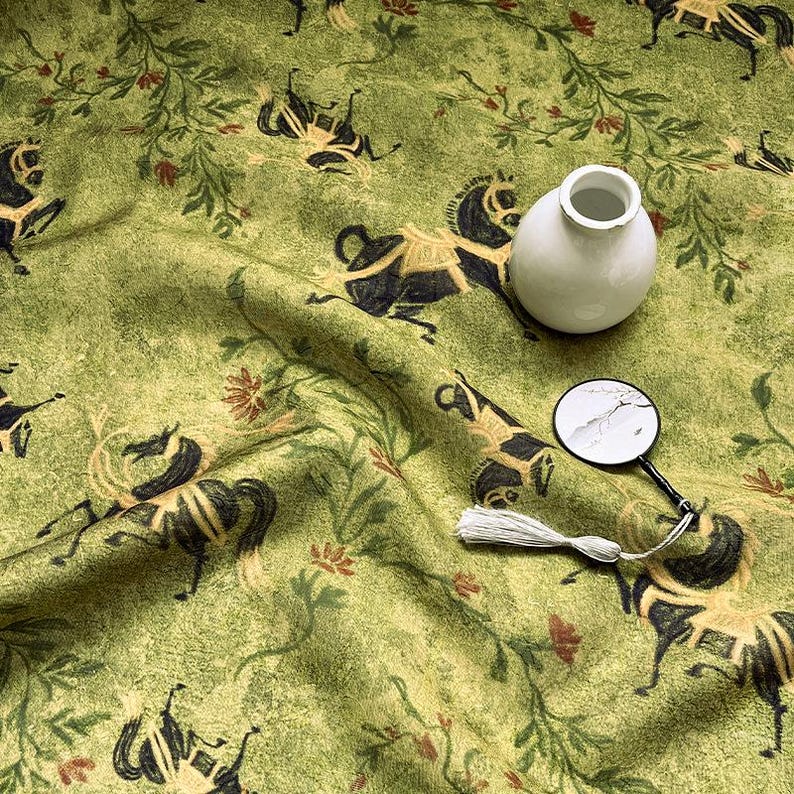 Może przedstawiać: Zbliżenie zielonej tkaniny z powtarzającym się wzorem czarnych koni i element&oacute;w kwiatowych. Na tkaninie znajduje się mały biały wazon i lupa z białą frędzlą.