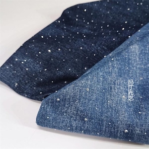 May include: Two sections of dark blue denim fabric, each adorned with scattered, sparkling embellishments. One piece is folded over the other. The corner of the lower piece displays white text. The denim has a textured appearance.