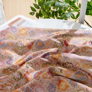 May include: Detailed shot of a patterned fabric featuring a repeating design of stylized animals and floral motifs in pink, orange, green, and purple. The fabric has a subtle sheen and a textured look, possibly a luxurious material.