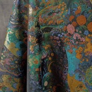 Puede incluir: Una tela vibrante con un dise&ntilde;o floral y paisaj&iacute;stico detallado. El patr&oacute;n presenta una mezcla de colores que incluyen el verde azulado, el naranja, el dorado y el negro. La tela est&aacute; drapeada, mostrando la intrincada obra de arte y la textura.