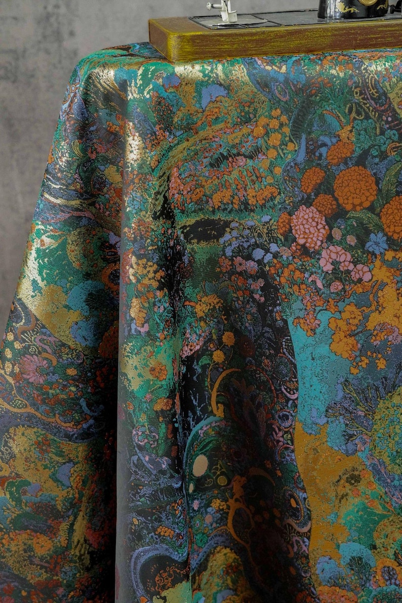 Puede incluir: Una tela vibrante con un dise&ntilde;o floral y paisaj&iacute;stico detallado. El patr&oacute;n presenta una mezcla de colores que incluyen el verde azulado, el naranja, el dorado y el negro. La tela est&aacute; drapeada, mostrando la intrincada obra de arte y la textura.