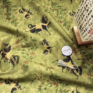 Może przedstawiać: Zielona tkanina z powtarzającym się wzorem czarnych koni ze złotymi akcentami i elementami kwiatowymi. Tkanina ma lekko pomarszczoną fakturę. Widoczny jest r&oacute;wnież mały dekoracyjny wachlarz i lampa z azjatyckimi znakami.