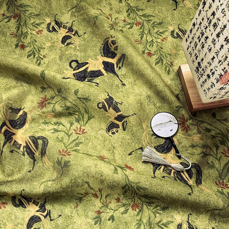 Może przedstawiać: Zielona tkanina z powtarzającym się wzorem czarnych koni ze złotymi akcentami i elementami kwiatowymi. Tkanina ma lekko pomarszczoną fakturę. Widoczny jest r&oacute;wnież mały dekoracyjny wachlarz i lampa z azjatyckimi znakami.