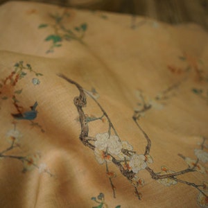 May include: A close-up of a beige fabric with a delicate floral print. The fabric features white flowers, green leaves, and a small blue bird. The fabric has a soft, flowing texture.