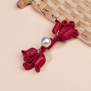 Könnte beinhalten: Eine weinrote Stoffblume mit einer großen Perle in der Mitte und kleinen goldfarbenen Akzenten. Die Blume hat ein geschichtetes Blütenblattdesign und ist wahrscheinlich eine dekorative Verzierung für Kleidung oder Accessoires.