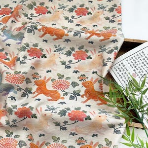 Puede incluir: Tela blanquecina con un patrón repetido de conejos, flores y follaje en tonos naranja, melocotón y verde. El diseño presenta conejos estilizados y elementos florales, creando un textil caprichoso y decorativo.